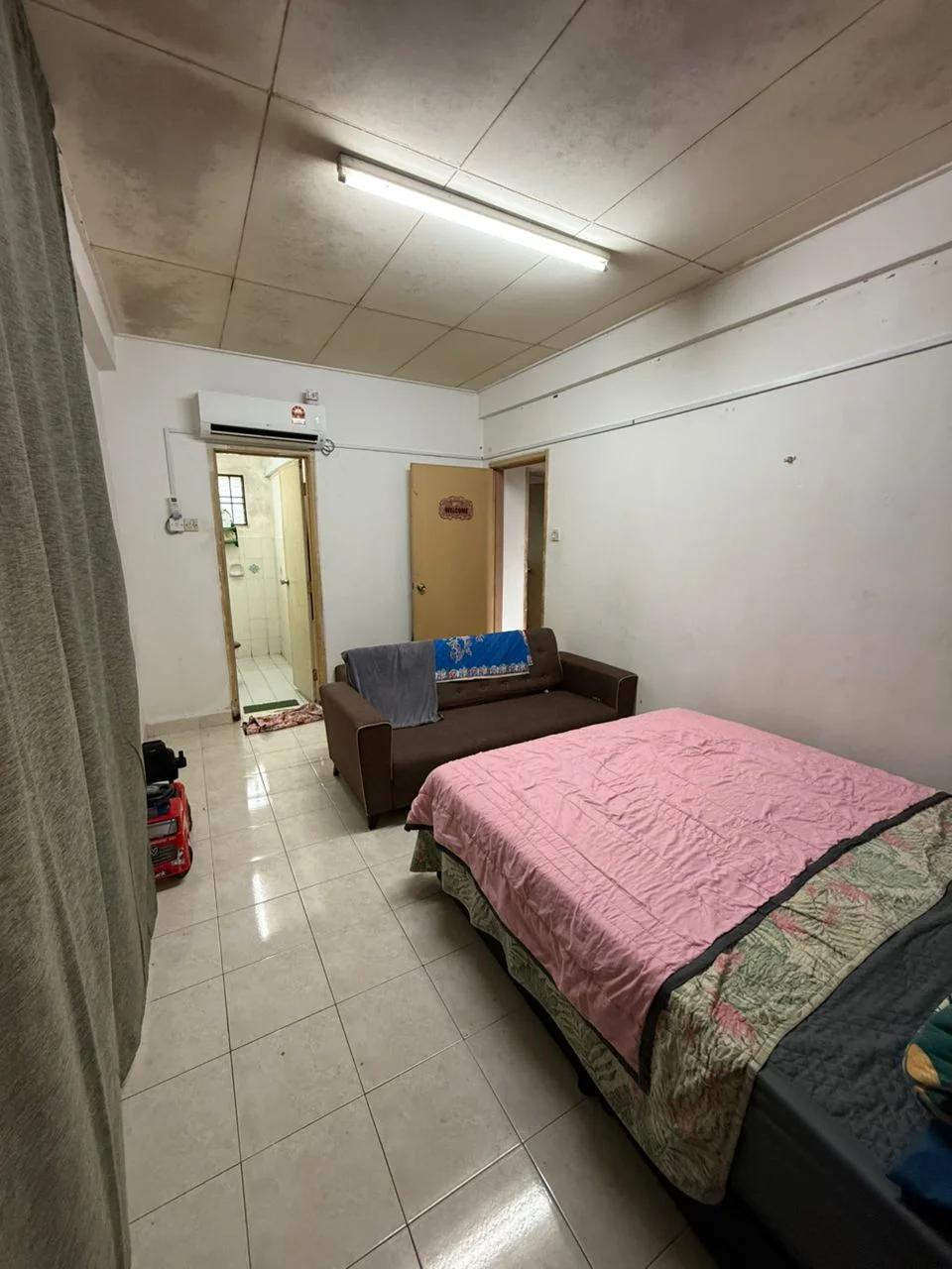 Room features: white painted walls, tile flooring, fluorescent ceiling lights, single bed with pink bedspread, brown fabric sofa, door leading to bathroom, window with sheer curtain, small area rug, wall-mounted air conditioning unit.